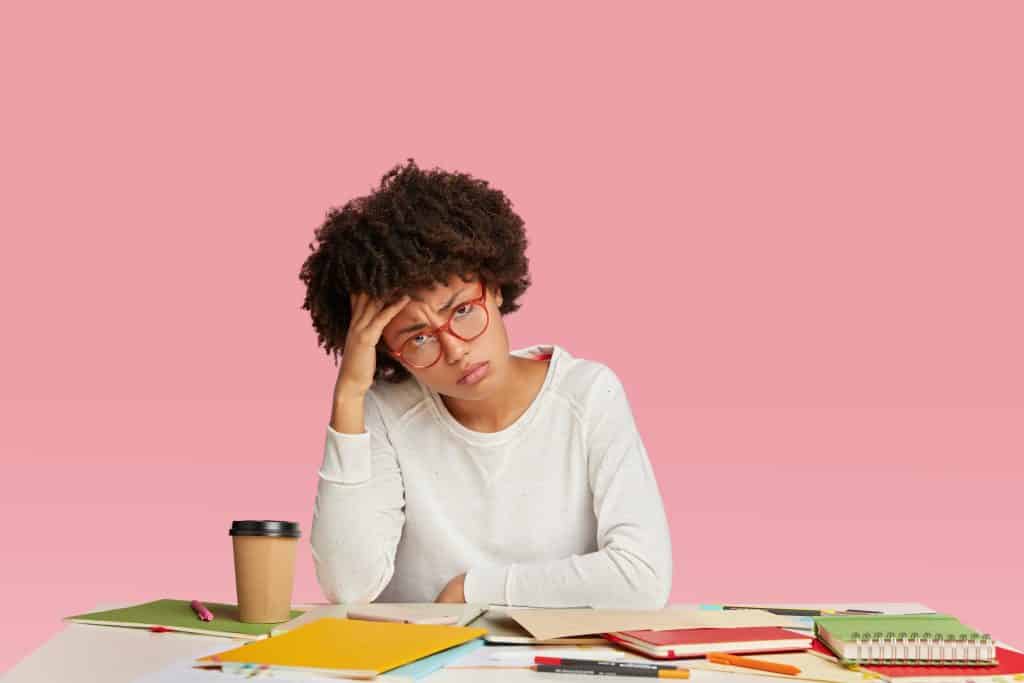 Tired dark skinned stressed businesswoman feels headache, looks fatigue, keeps hand on head, wears white casual sweater, spectacles with red rim, isolated over pink background, busy with work